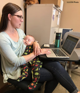 Angie working at her laptop, sitting, while holding her sleeping baby