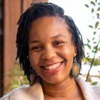 Black person with short hair, in braids, and smile, teeth showing, is looking at the camera.